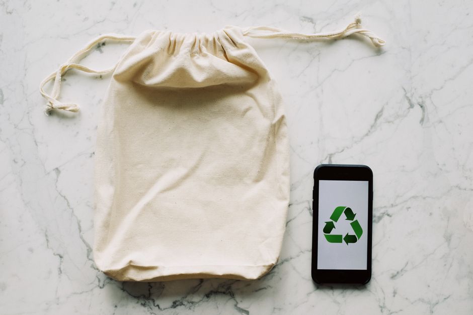 A beige, reusable fabric drawstring bag with a tied top, placed on a white marble surface with grey veining. To the right of the bag, there is a black smartphone displaying a green recycling symbol on