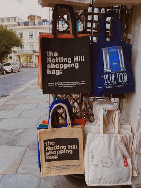 A beige, reusable fabric drawstring bag with a tied top, placed on a white marble surface with grey veining. To the right of the bag, there is a black smartphone displaying a green recycling symbol on
